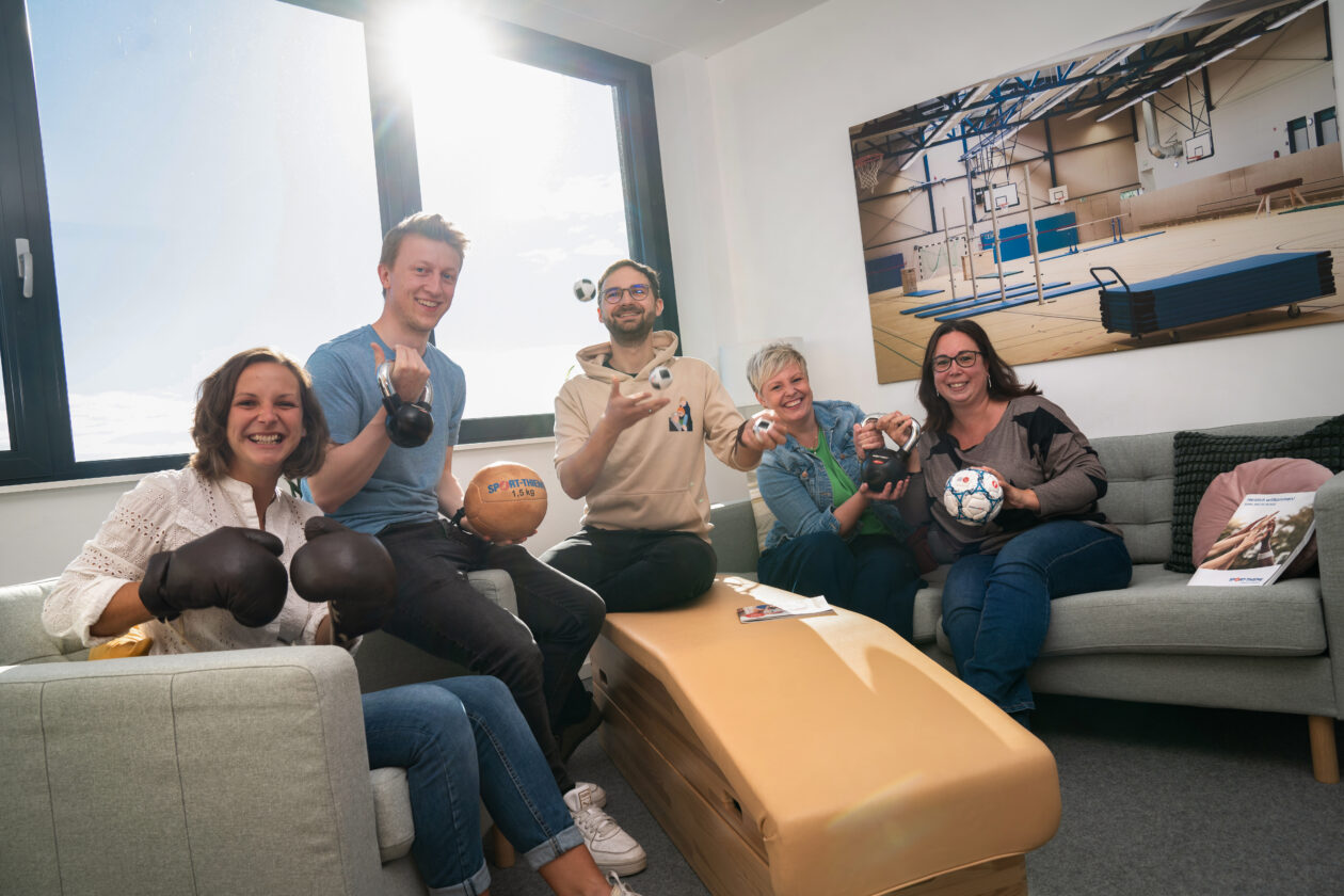 Five employees from Sport-Thieme's HR department are sitting in a meeting room holding various pieces of sports equipment.