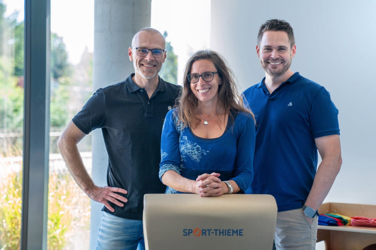Die Geschäftsleitung von Sport-Thieme mit Maximilian Hohe, Katharina Hohe-Thieme und Michael Egerer steht gemeinsam vor einer Sport-Thieme-Bank und blickt freundlich in die Kamera.
