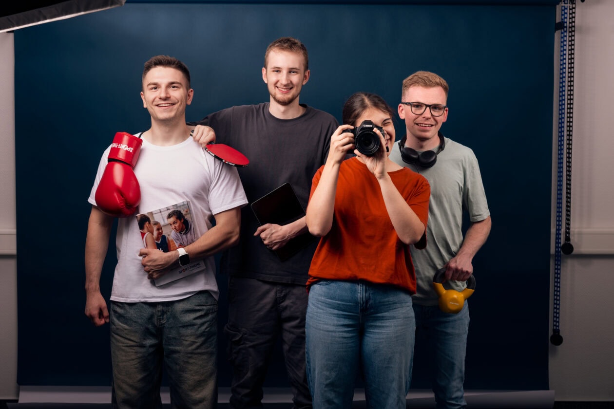 Four Sport-Thieme trainees standing together in front of a dark blue backdrop. One person is holding red boxing gloves and a catalog, another holds a tablet. The person in the orange shirt is taking a photo with a camera, while the fourth person holds a yellow kettlebell.