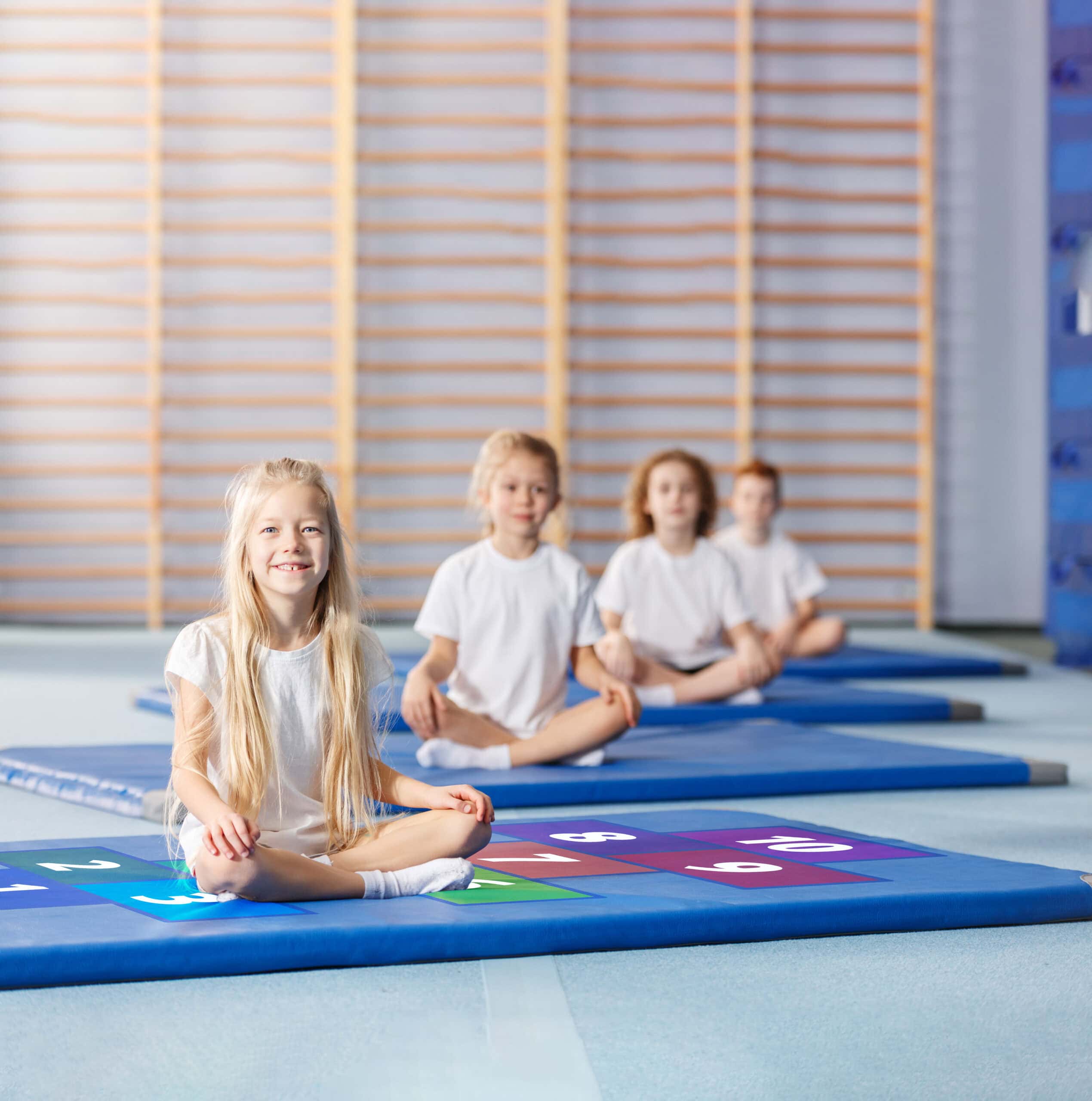 Kinder sitzen auf blauen Turnmatten in einer Sporthalle, teilweise auf Matten mit bunten Zahlenfeldern.